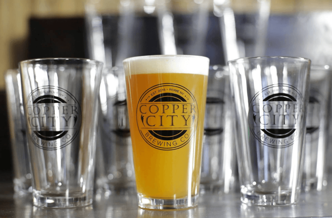 A pint glass filled with hazy beer sits among empty Copper City Brewing glasses, their logos visible. The background is softly blurred, drawing attention to the full glass in the center.